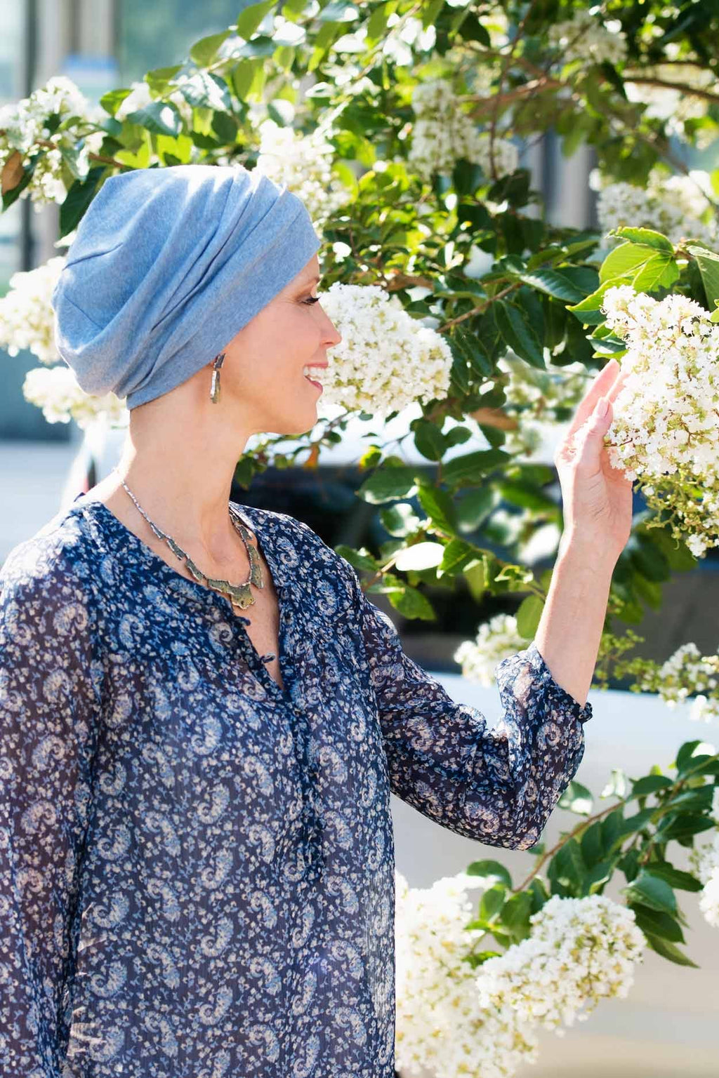 Woman outdoors with white flowers wearing denim blue colored chemo beanie hat