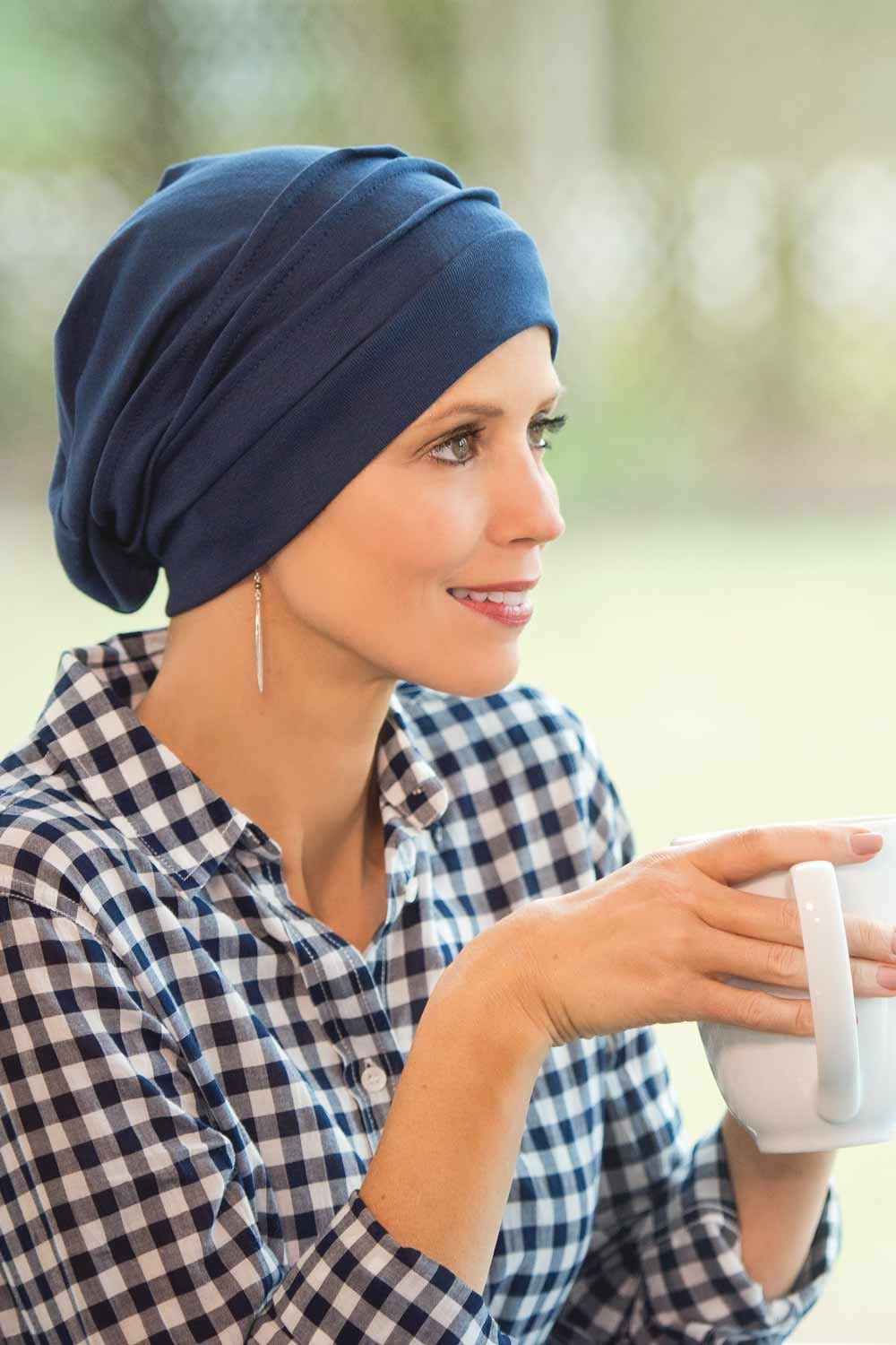 Woman drinking coffee by window wearing blue slouchy snood hat designed for cancer patients